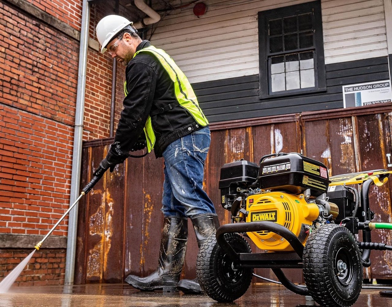 Worker in a hard hat and safety vest pressure washing a paved area beside a brick building